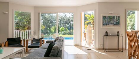 Una sala de estar moderna con un sofá, una mesa de comedor y sillas. Se ve un ventanal con vistas a una piscina y follaje.