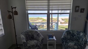 Una habitación con dos sillones estampados, una mesita con un libro, una lámpara de pie y vistas a la playa a través de una ventana.