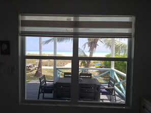 Ein Zimmer mit großem Fenster mit Blick auf einen Strand, einer Holzterrasse mit Gartenmöbeln und Meerblick.