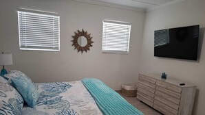 Un dormitorio con cama, dos ventanas con persianas, un televisor montado en la pared, un aparador y un espejo.