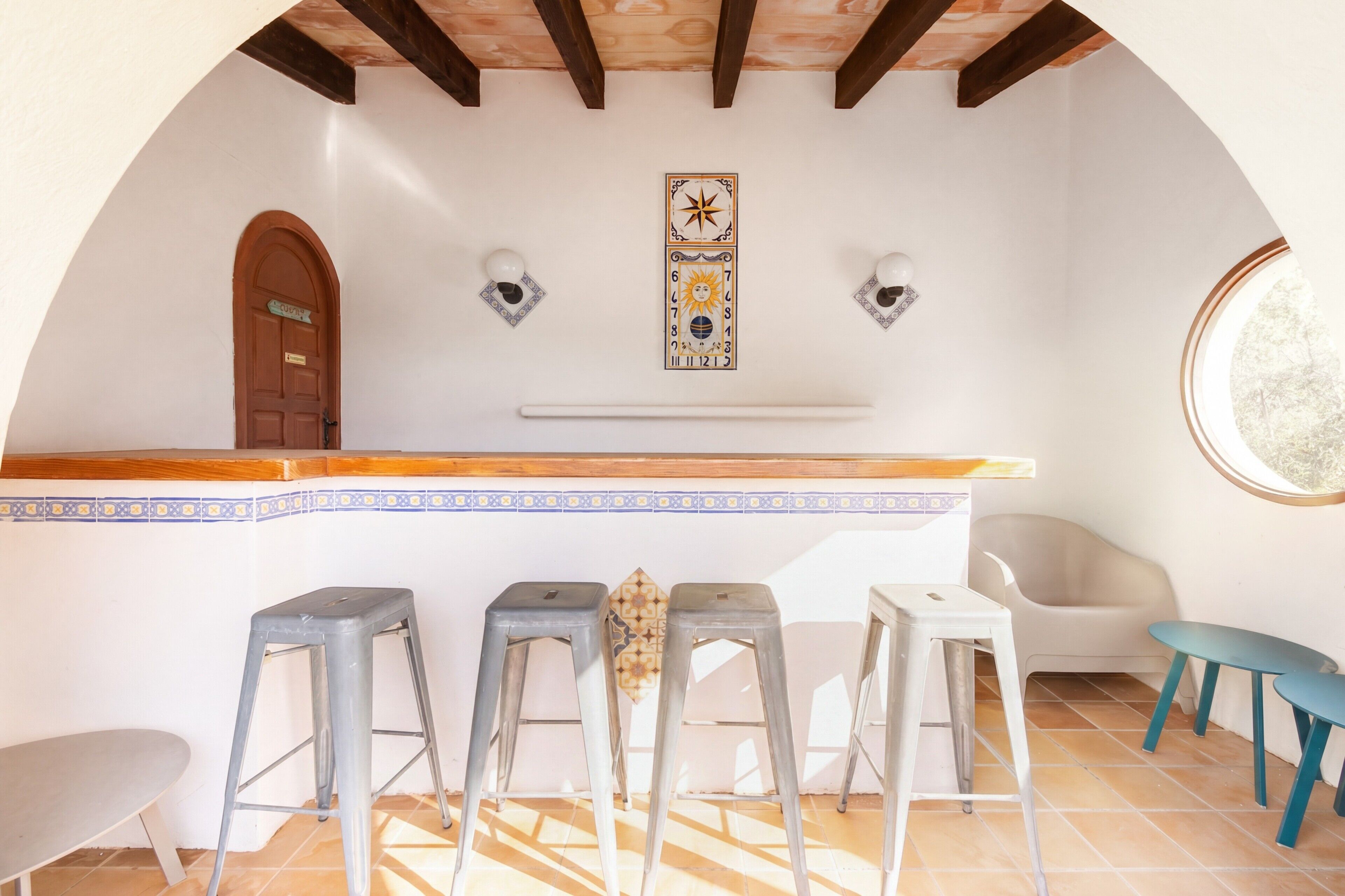 Un techo abovedado con vigas de madera, una zona de bar con taburetes, una puerta y apliques de luz en la pared.