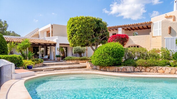 A poolside view of a resort with a clear pool, a covered seating area, and a well-maintained garden.