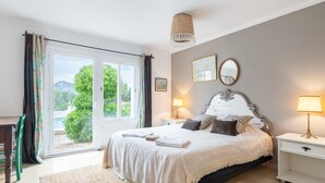 A bedroom with a bed, two bedside tables, a hanging light fixture, a window with curtains, and a view of greenery outside.