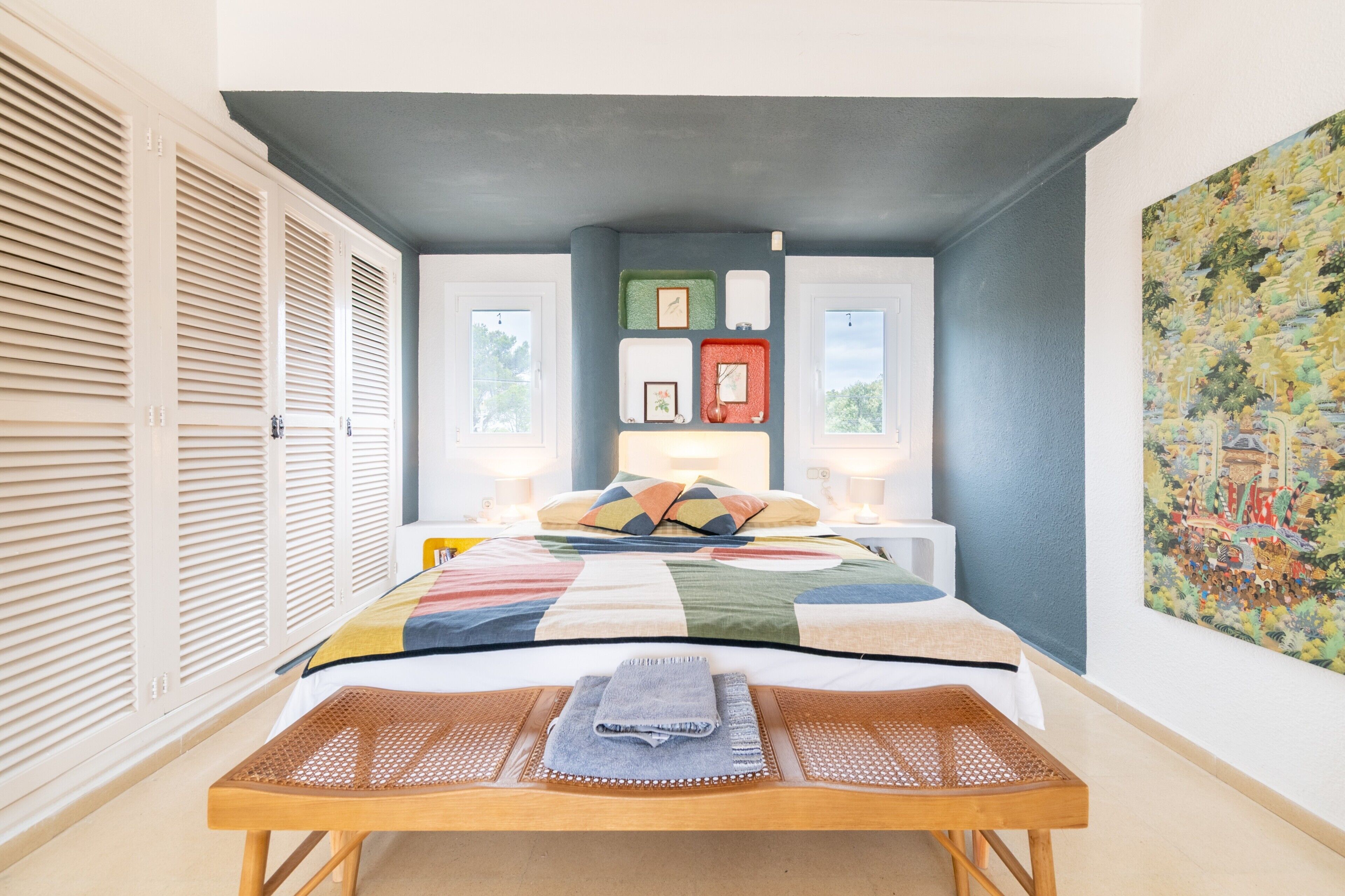 Un dormitorio con cama, mesitas de noche, ventana y un cuadro en la pared.