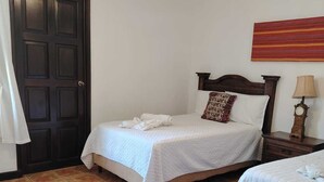 A bedroom with two beds, a wooden door, a clock on a bedside table, and a wall hanging. - Alcázar de San Bartolo (Antigua Guatemala)