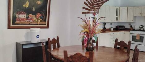 A dining area with a round wooden table and chairs, a painting on the wall, and a kitchen in the background.
