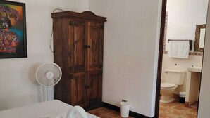 Ein Schlafzimmer mit Bett, einem Holzschrank, einem weißen Ventilator und einem durch eine offene Tür einsehbaren Badezimmer.