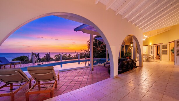 Uma área externa coberta com piscina, espreguiçadeiras e vista para o oceano ao pôr do sol.