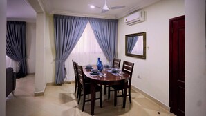 A dining area with a round table set for four, surrounded by chairs, with a ceiling fan above and an air conditioning unit on the wall. - Willows Court (Accra)