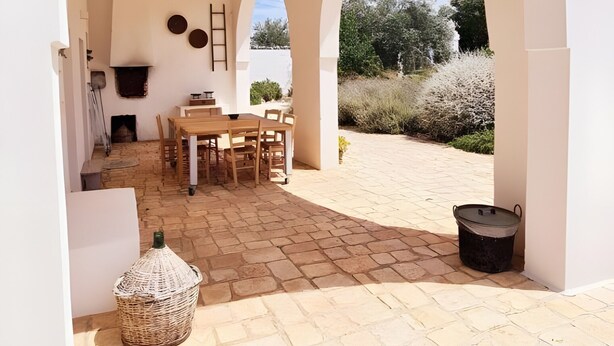 An outdoor patio with a stone floor, a wooden table and chairs, and a white wall with a fireplace. - Apartment 'Masseria San Martino -3' with Shared Pool, Wi-Fi and Air Conditioning (Polignano a Mare)