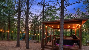Un gazebo in legno con veranda e area salotto, circondato da alberi e da un’area giochi.
