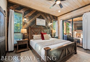A bedroom with a wooden headboard, a bed with patterned bedding, a nightstand with a lamp, and a view of an outdoor area. - Tender Retreat in the Pines | Game-Filled Retreat (Broken Bow)