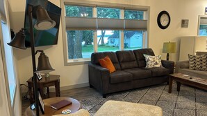 Ein Wohnzimmer mit einem dunkelbraunen Sofa, einem runden Couchtisch, einer Stehlampe, einem Fernseher und einem Fenster mit Jalousien.