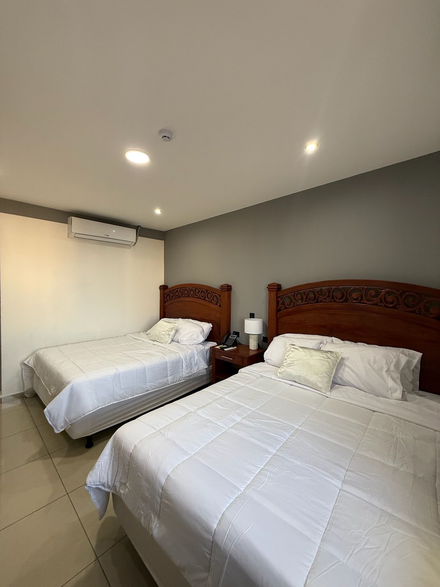A hotel room with two beds, a bedside table with a lamp, and an air conditioning unit on the wall.