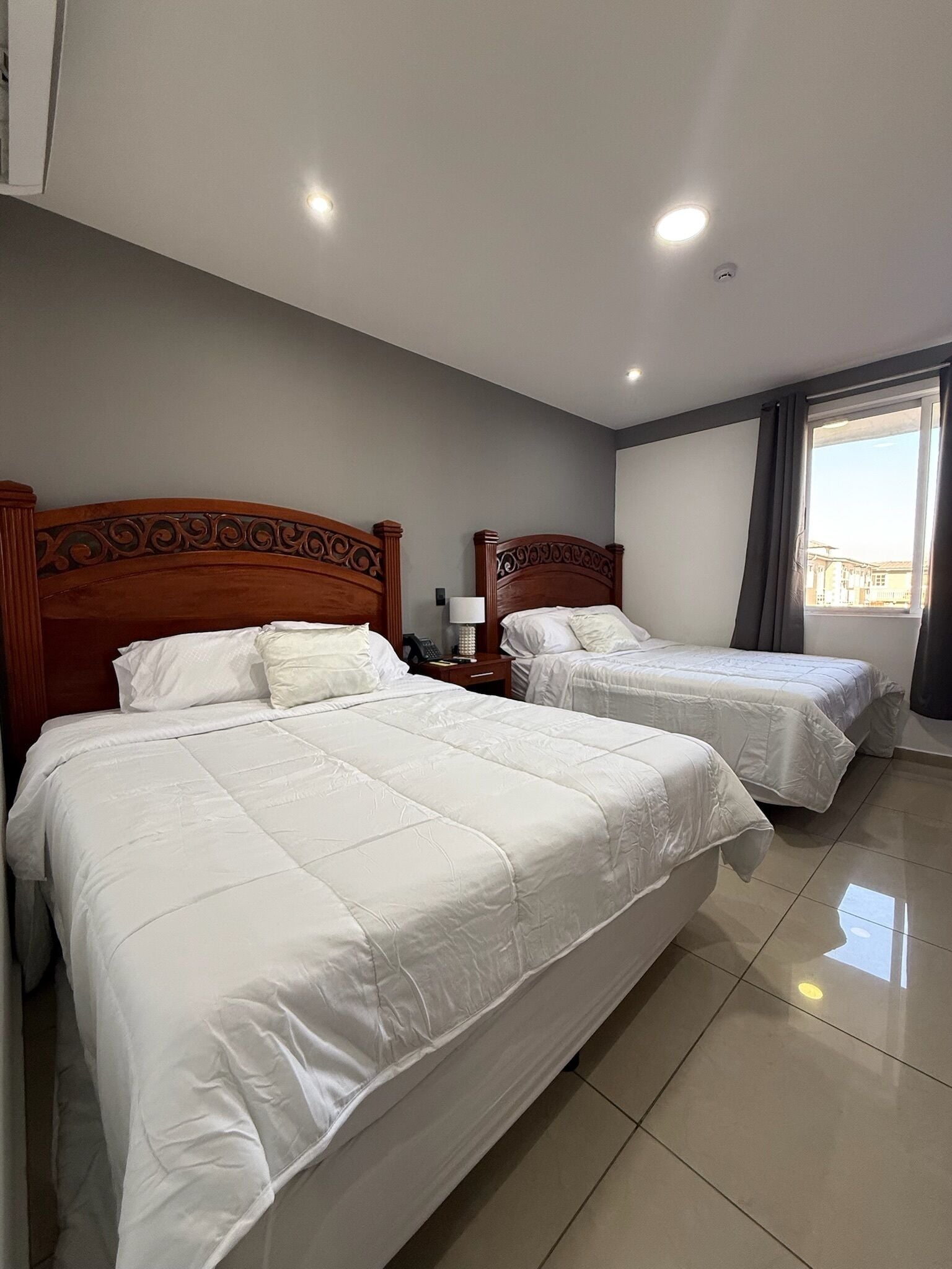 A hotel room with two beds, a nightstand, a lamp, and a window with curtains.