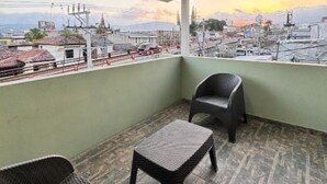 Un balcon donnant sur un paysage urbain, une chaise en osier, une petite table et une rambarde.