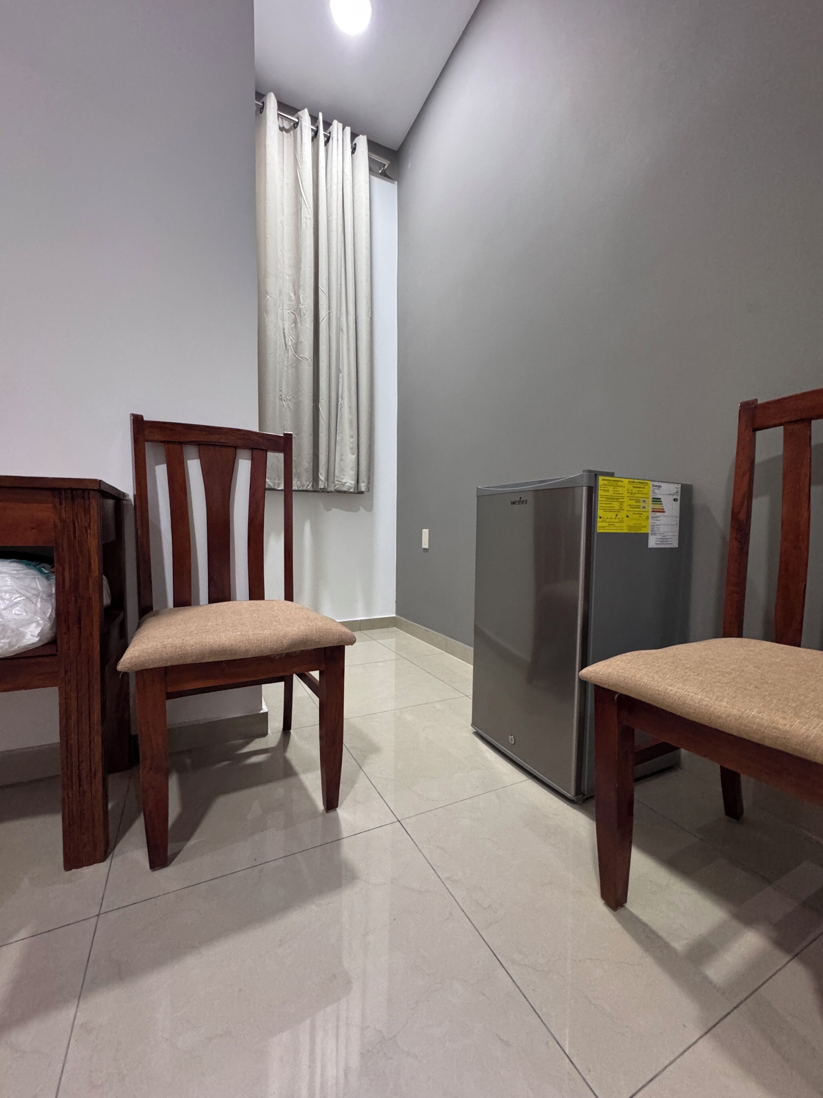 A room with two wooden chairs, a refrigerator, a window with curtains, and a tiled floor.