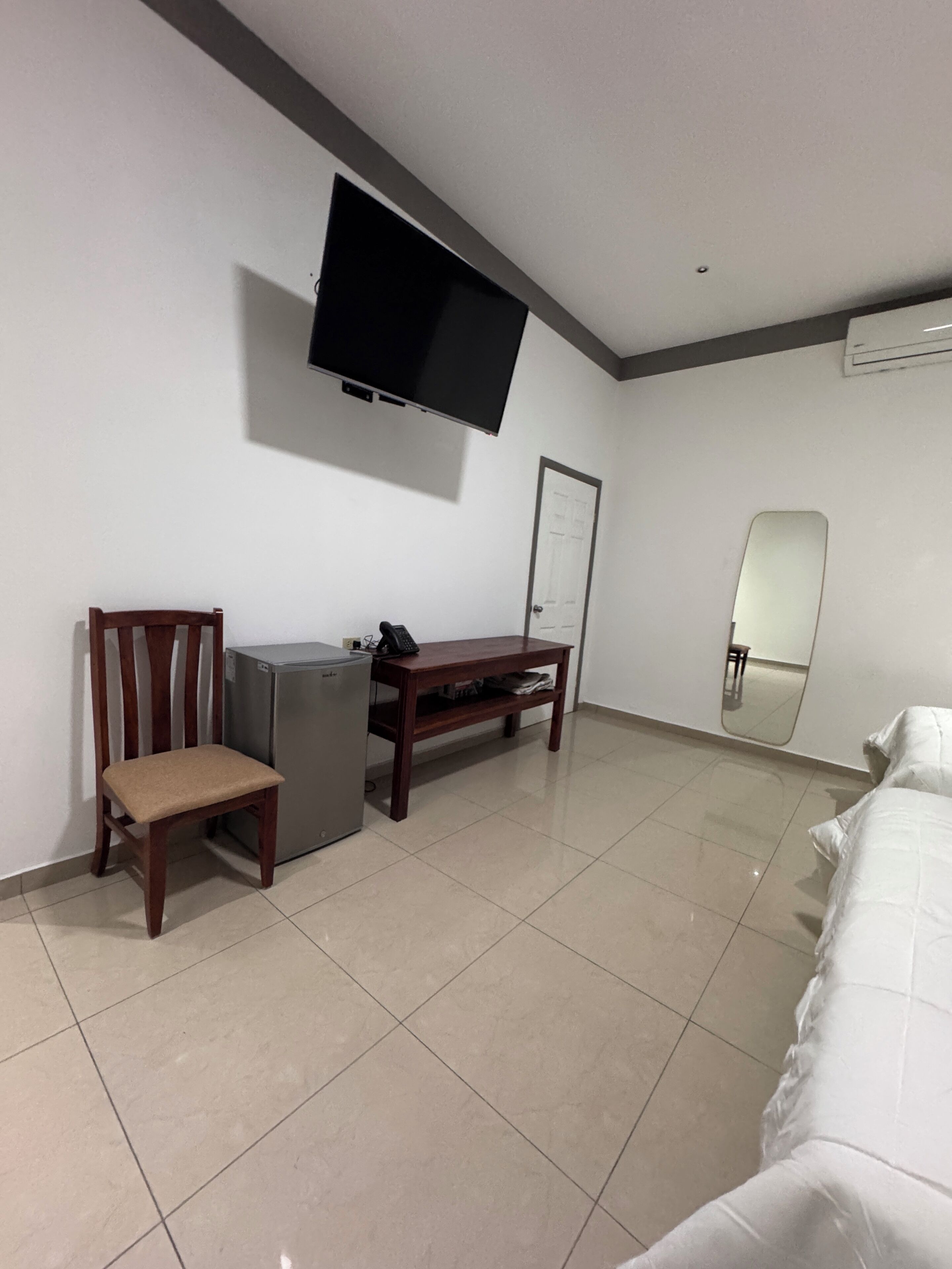 A hotel room with a bed, a TV mounted on the wall, a small desk, a chair, and a refrigerator.