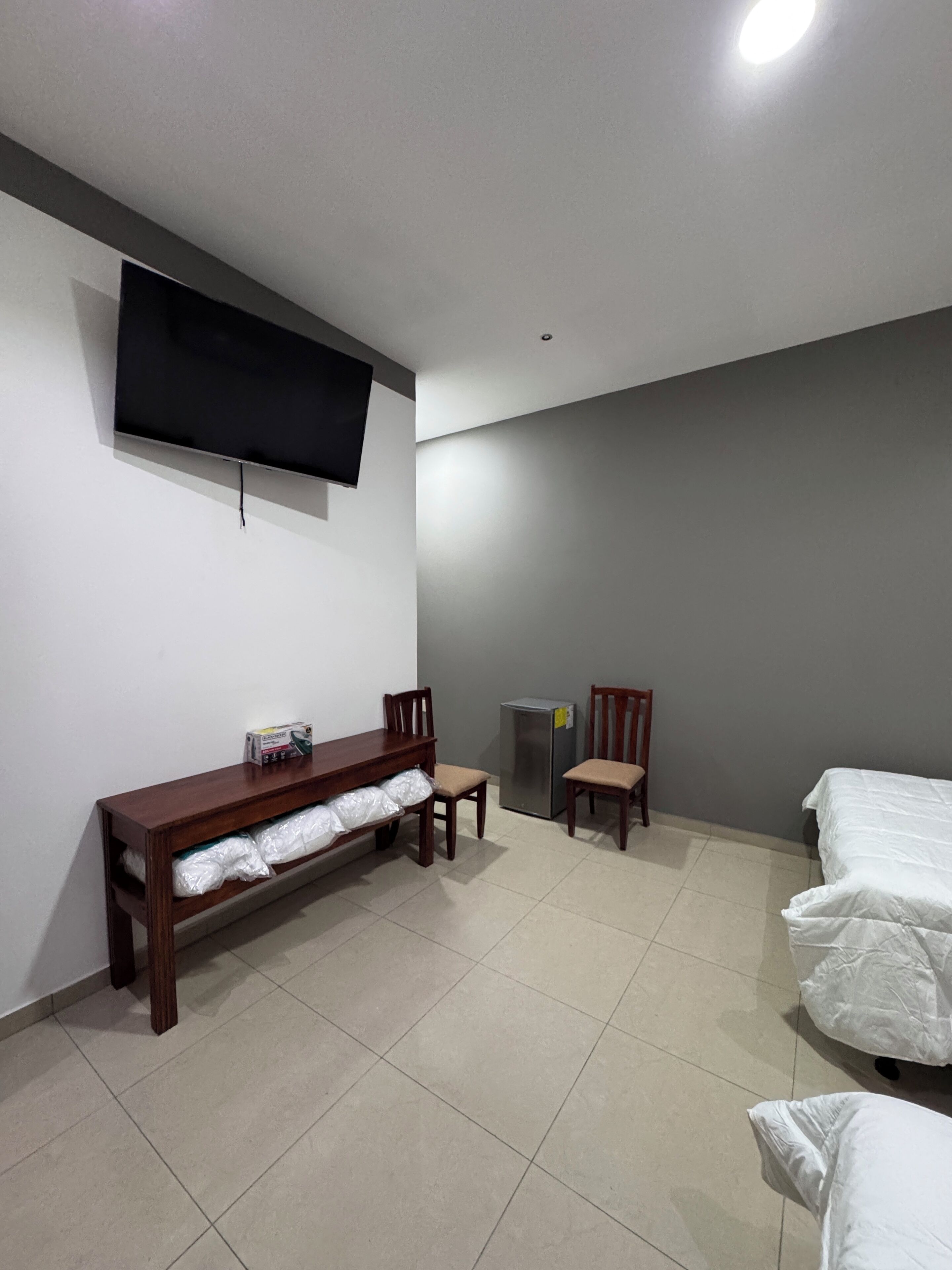 Une pièce avec un téléviseur fixé au mur, un banc en bois avec espace de rangement, deux chaises, un lit et un réfrigérateur.