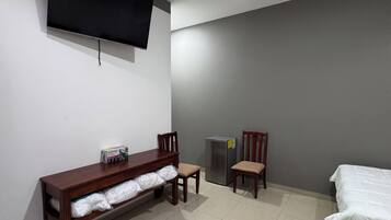 Une pièce avec un téléviseur fixé au mur, un banc en bois avec espace de rangement, deux chaises, un lit et un réfrigérateur.