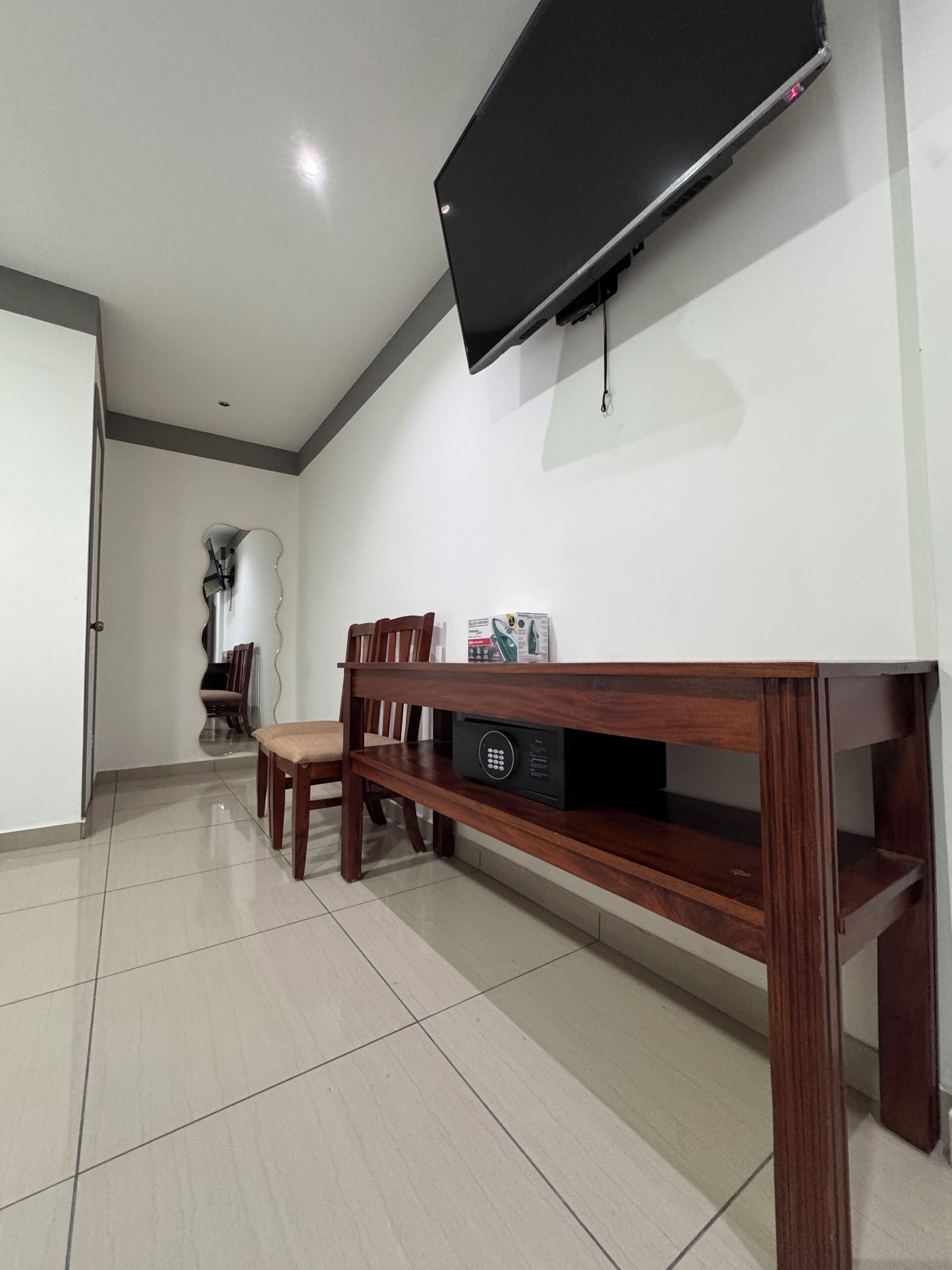 A room with a flat-screen TV mounted on the wall, a wooden dining table with chairs, and a tiled floor.