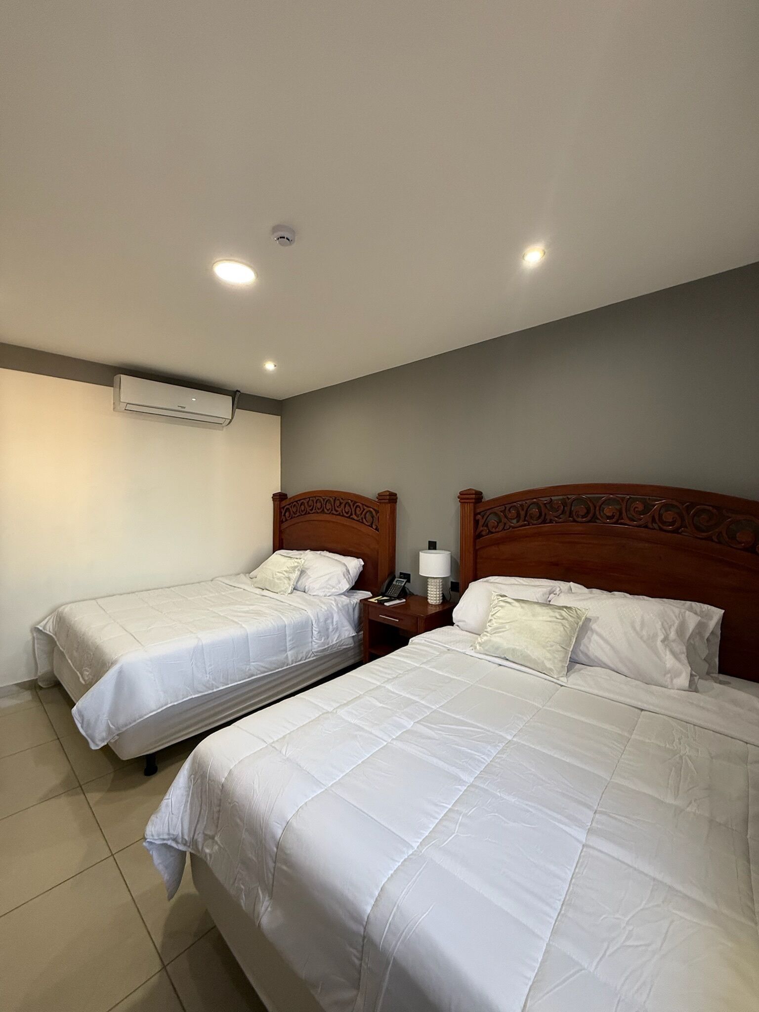 A hotel room with two beds, a ceiling-mounted air conditioner, a bedside table with a lamp, and a phone.