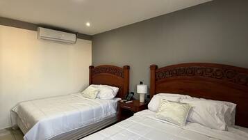 A hotel room with two beds, a ceiling-mounted air conditioner, a bedside table with a lamp, and a phone.