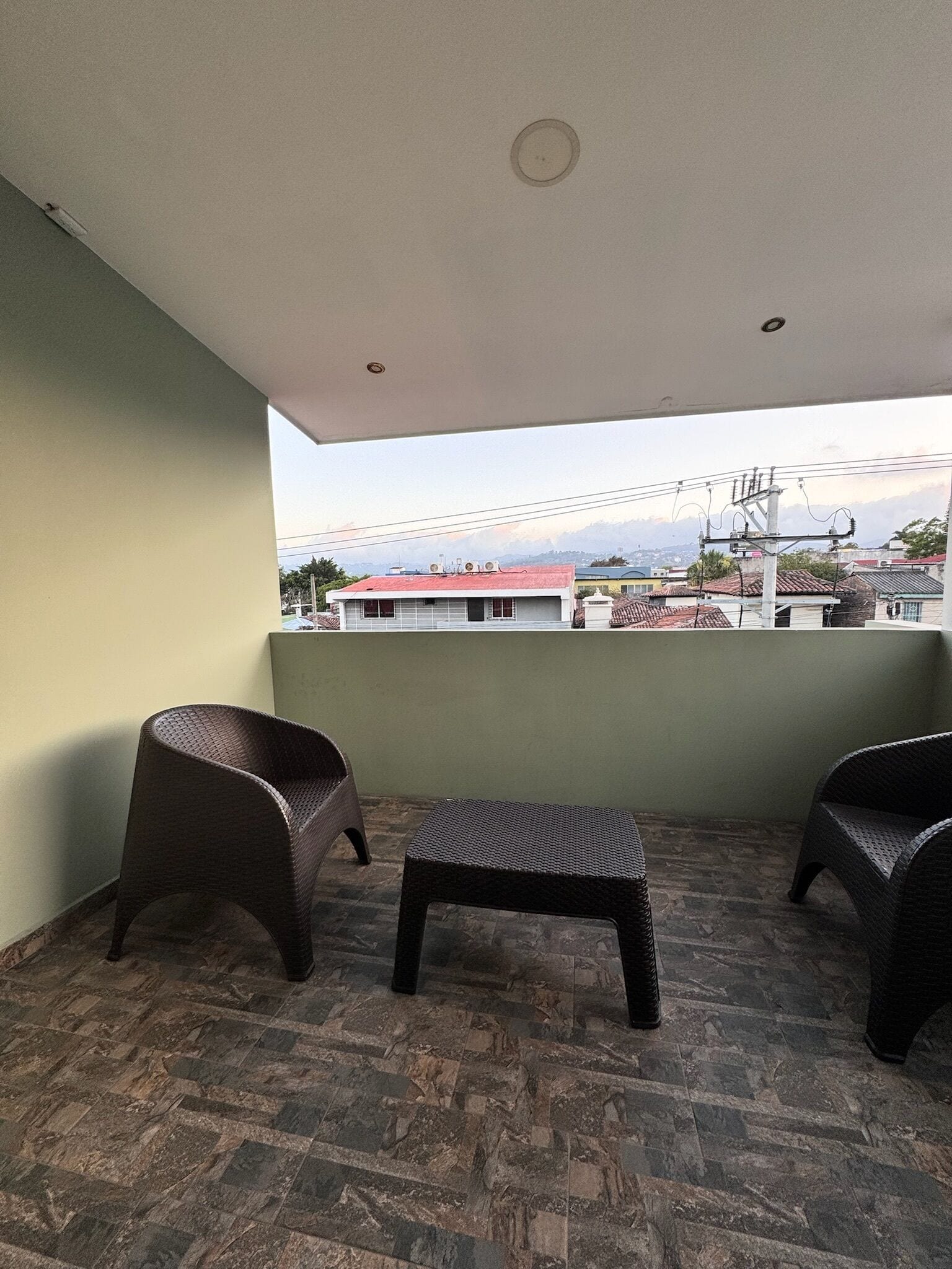 Un balcón con dos sillas de mimbre y un pequeño reposapiés, suelo de baldosas y vista a edificios y líneas eléctricas al fondo.