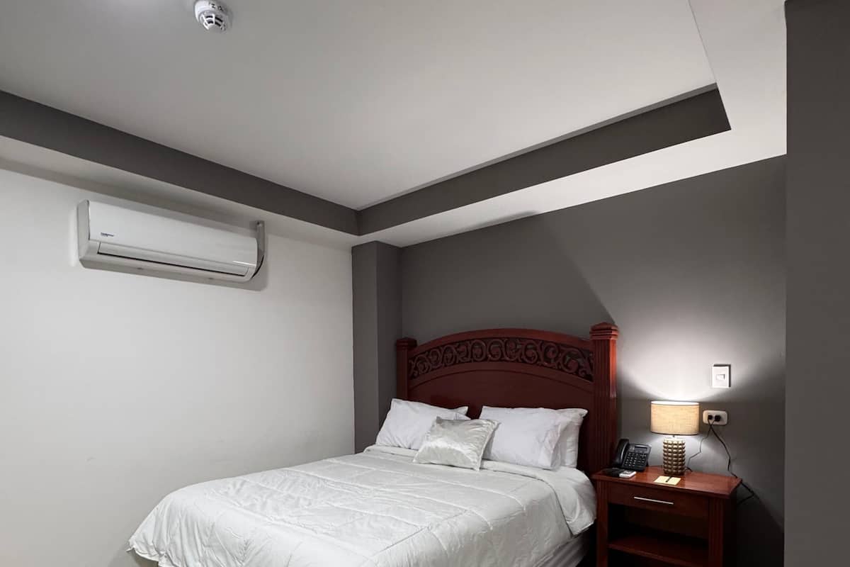 Una cama bien hecha con ropa blanca, cabecera de madera, mesita de noche con lámpara y un equipo de aire acondicionado en la pared.