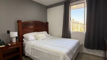 A hotel room with a bed, bedside table, lamp, and a window with curtains.