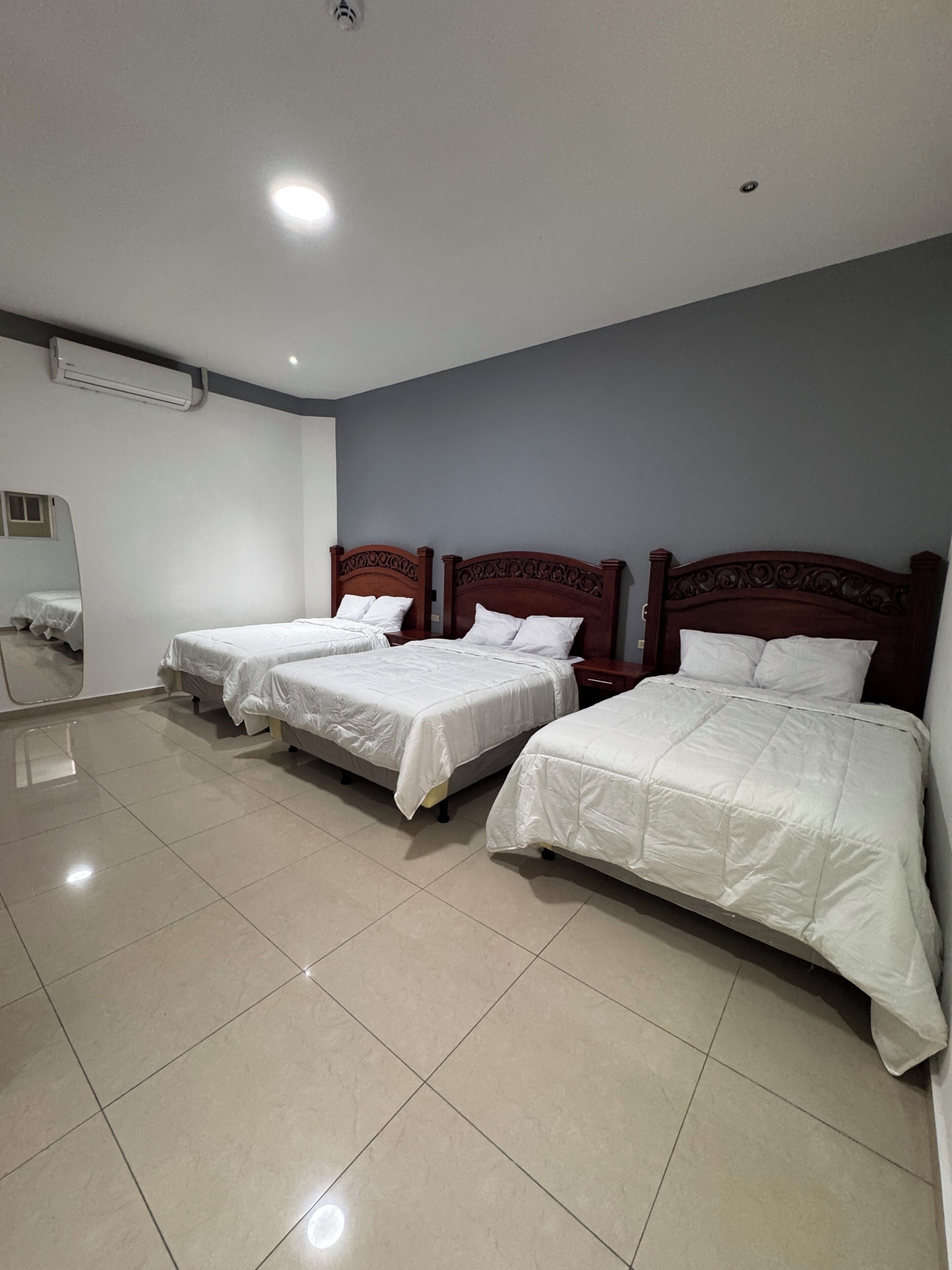 Une chambre d’hôtel avec trois lits simples, un grand miroir et un sol carrelé.
