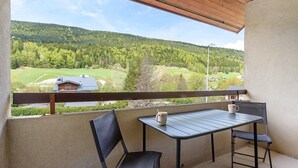 A balcony with a table and chairs, a view of a green landscape, and a house in the distance. - Elegant 'Havre de Paix' Apartment with Mountain View, Private Terrace & Wi-Fi (Villard-de-Lans)
