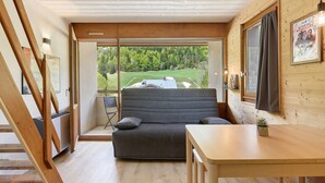 A modern interior with a sofa, a dining table, and a view of a green landscape through large windows. - Elegant 'Havre de Paix' Apartment with Mountain View, Private Terrace & Wi-Fi (Villard-de-Lans)