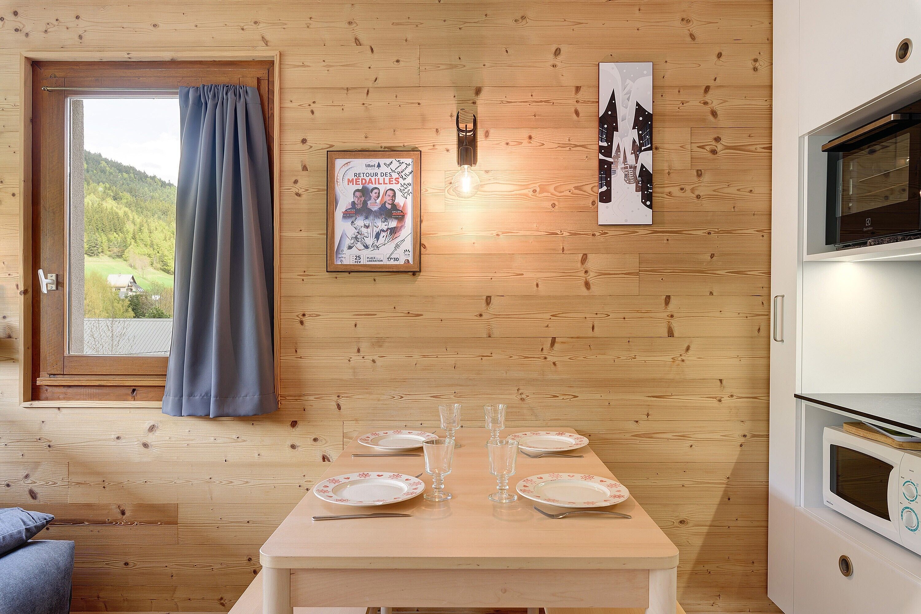 Une pièce aux murs lambrissés, avec une table à manger dressée pour quatre personnes, une fenêtre avec des rideaux, une affiche accrochée au mur et un four à micro-ondes.