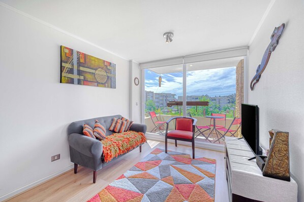Un salon moderne avec un canapé gris, un plaid coloré et un tapis à motifs géométriques. On peut voir une chaise rouge et un meuble pour la télévision.