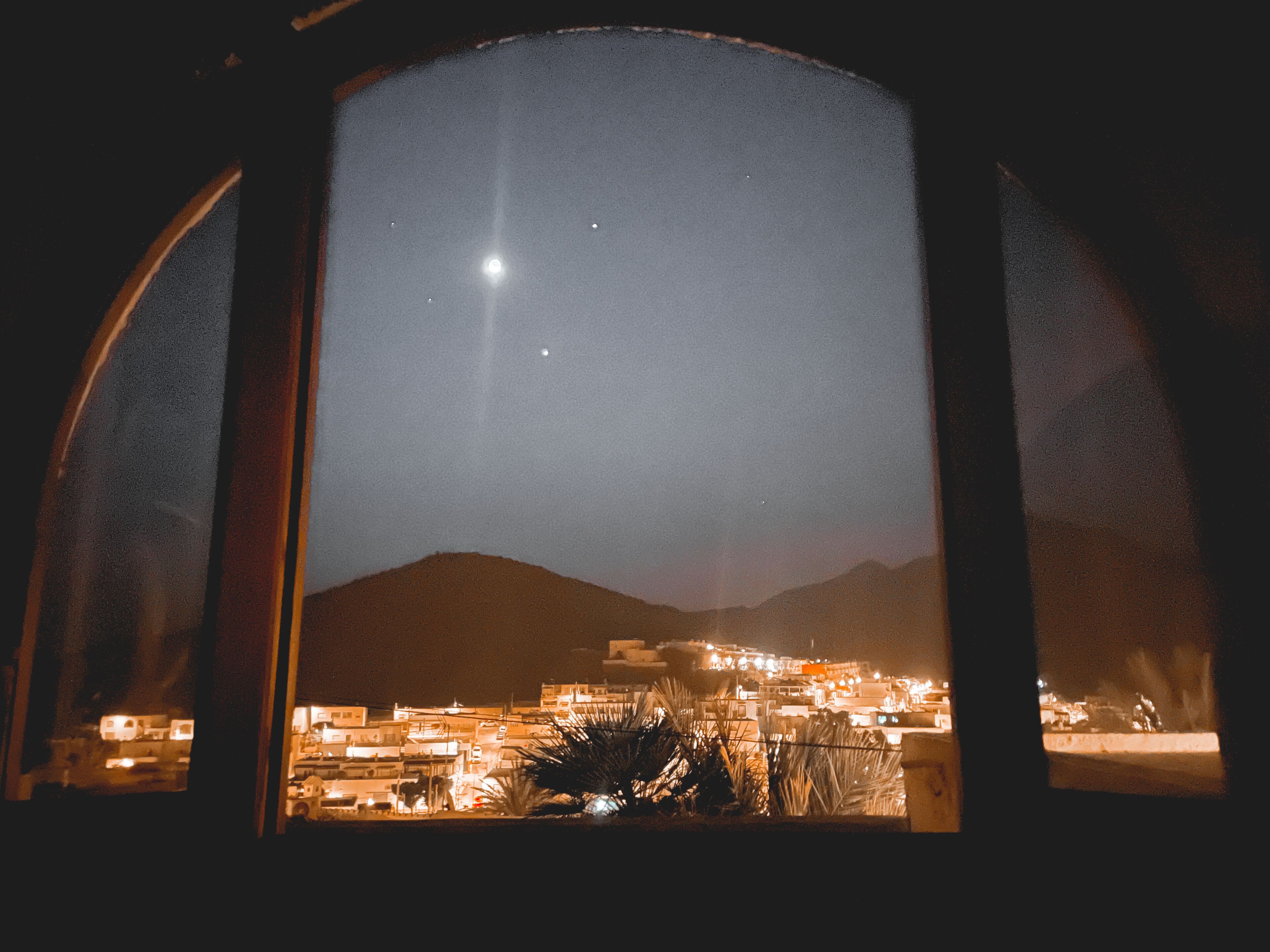 A nighttime view through a window, featuring a crescent moon, a star, and a cityscape with buildings and mountains in the background.