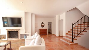 A spacious living room with a fireplace, white furniture, a TV, and a staircase. - Holiday Home 'Ses Gavines en Cala Moragues' with Sea View, Wi-Fi and Air Conditioning (Andratx)