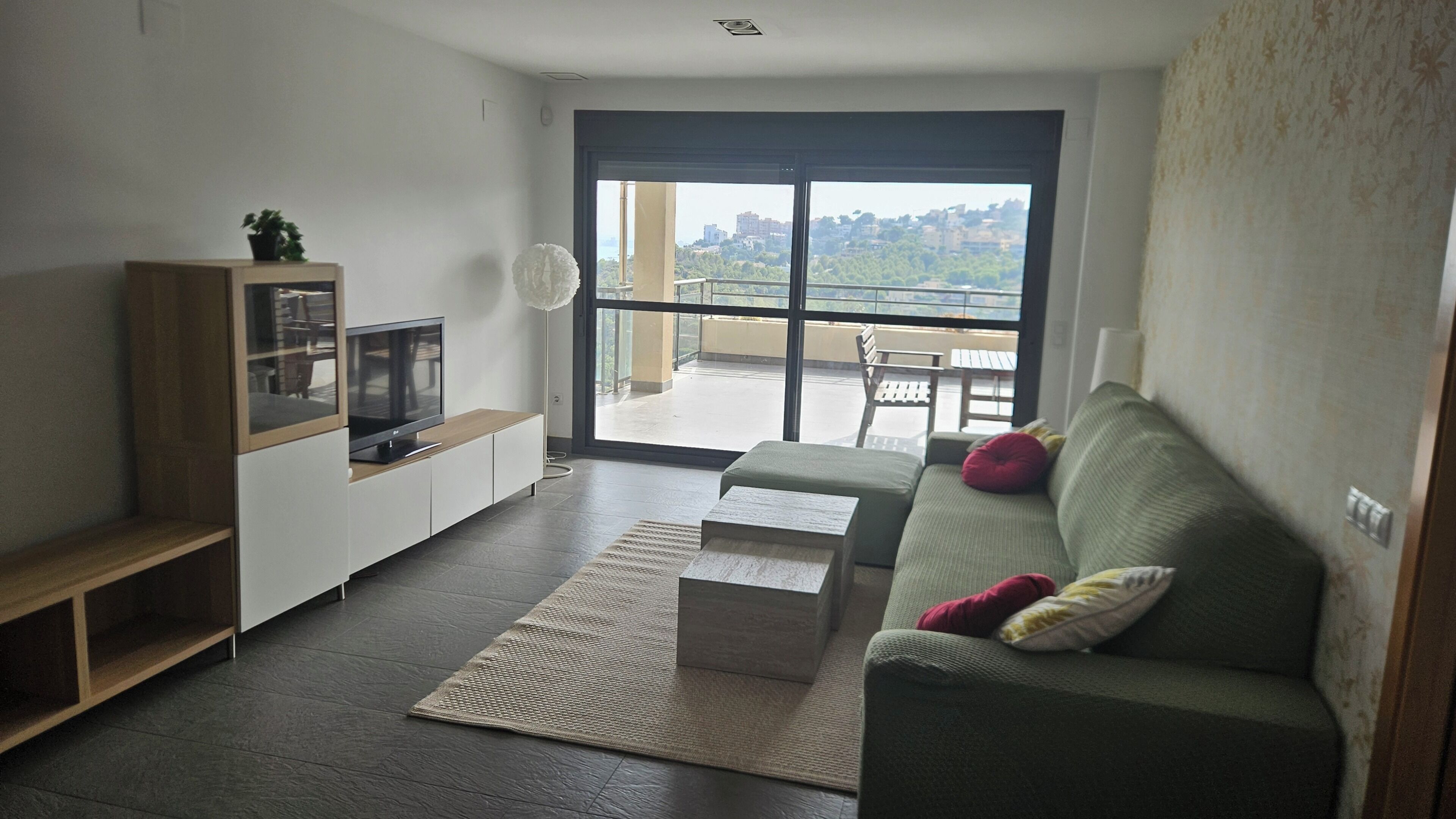 A modern living room with a flat-screen TV, a sofa with cushions, a coffee table, and a large sliding glass door leading to an outdoor balcony.