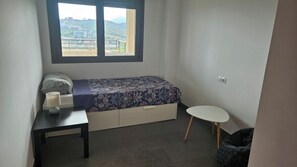 A small, tidy room with a bed, a desk, a chair, and a window with a view of buildings and hills. - Luxury apartment with sea view (VC)
