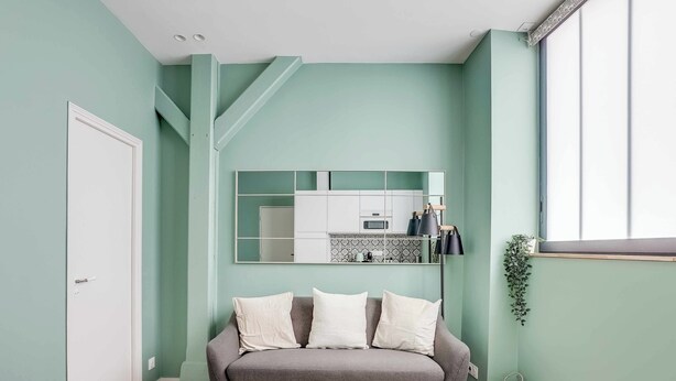 Una sala de estar con un sofá gris, una mesa de centro de vidrio, una zona de cocina con armarios blancos y un ventanal con una planta.