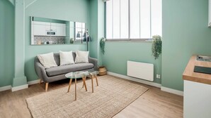 Una sala de estar moderna con un sofá gris, una mesita de centro, un ventanal con persianas, y una cocina visible a través del ventanal.