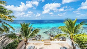 Vista de playa con agua turquesa cristalina, palmeras y un edificio blanco con escaleras que conducen a una zona de arena.