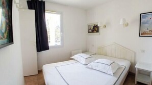 A single bed with white bedding and a headboard, a small bedside table, a window with black curtains, and a wall-mounted lamp. - Apartment with pools and garden near the beach (La Baule-Escoublac)