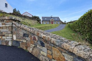 A stone wall leading to a house with a red door, a stone roof, and a clear sky.