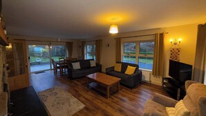 Un salon comprenant un canapé, une table basse et un téléviseur. Il y a également un coin repas avec des chaises et une table. De grandes fenêtres laissent entrer la lumière naturelle et offrent une vue sur l’extérieur.