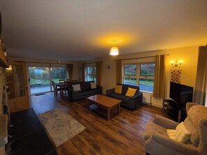 A living room with a sofa, coffee table, and TV. There is a dining area with chairs and a table. Large windows provide natural light and views of the outdoors.