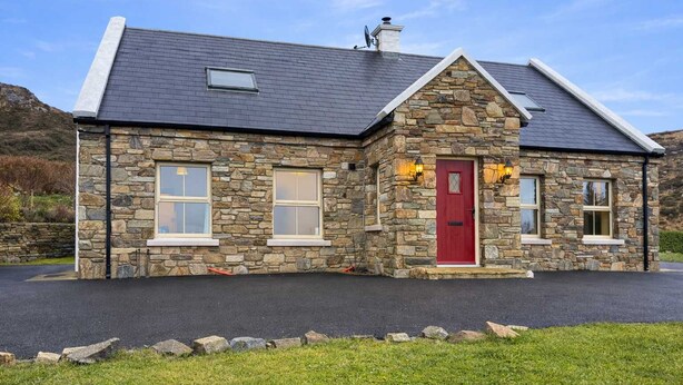 Une maison en pierre avec une porte rouge, deux fenêtres et une cheminée.