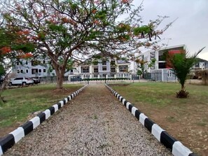 Uma trilha conduz a um edifício com estilo arquitetônico moderno, cercado por um gramado bem cuidado e uma árvore com folhas verdes e vermelhas.