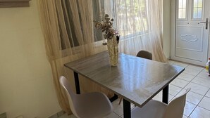 Zona pranzo con tavolo, due sedie, un vaso con fiori, una finestra con tende leggere e un cartello di uscita.