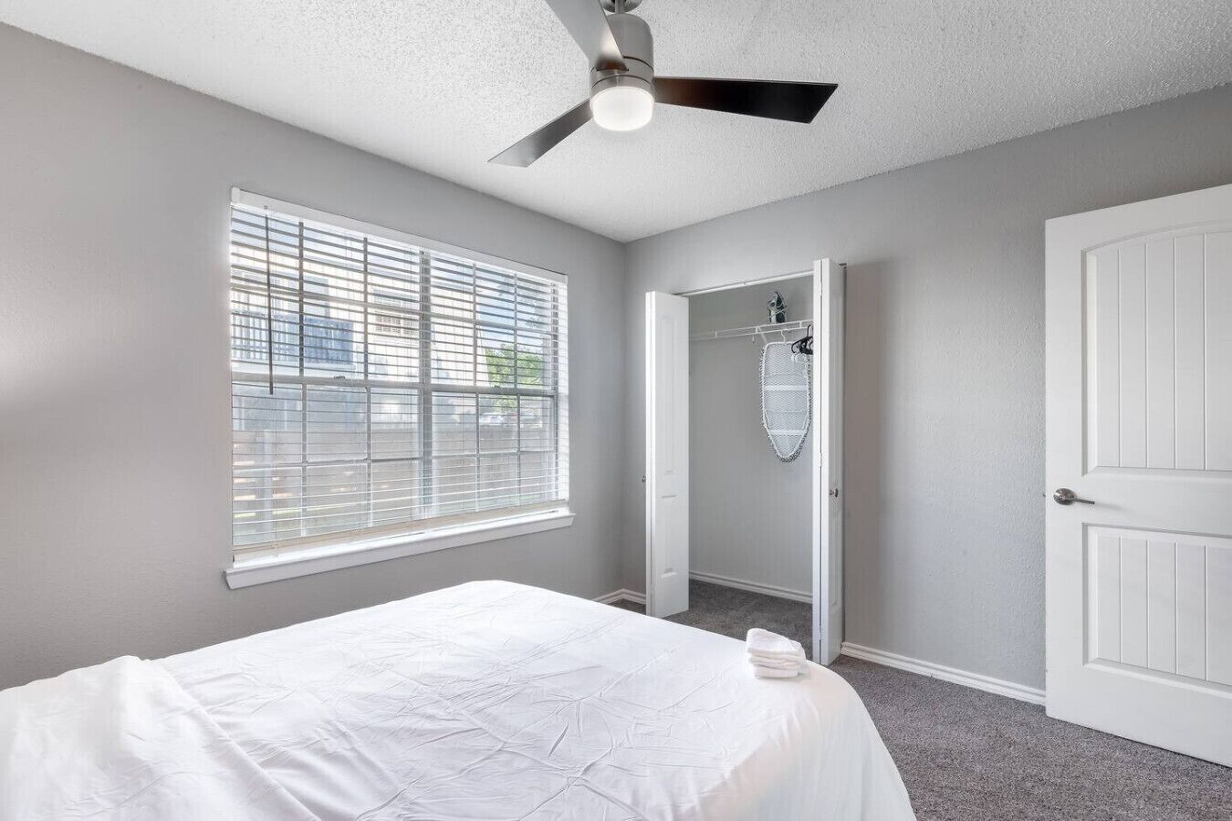 Une chambre à coucher comprenant un lit, un ventilateur de plafond, une fenêtre avec des stores, une porte et un dressing.